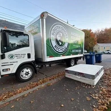 Green and white South Shore Recycling truck parked on a street beside a large mattress on the ground, with blue recycling bins nearby and a residential house in the background.