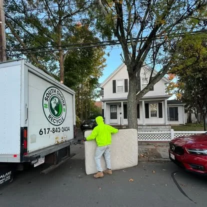 A person wearing a bright yellow-green hoodie and gray pants standing on a sidewalk, pushing a large foam or upholstered piece of furniture, in front of a large tree and residential house, with a South Shore Recycling truck parked nearby and a red car on the street.