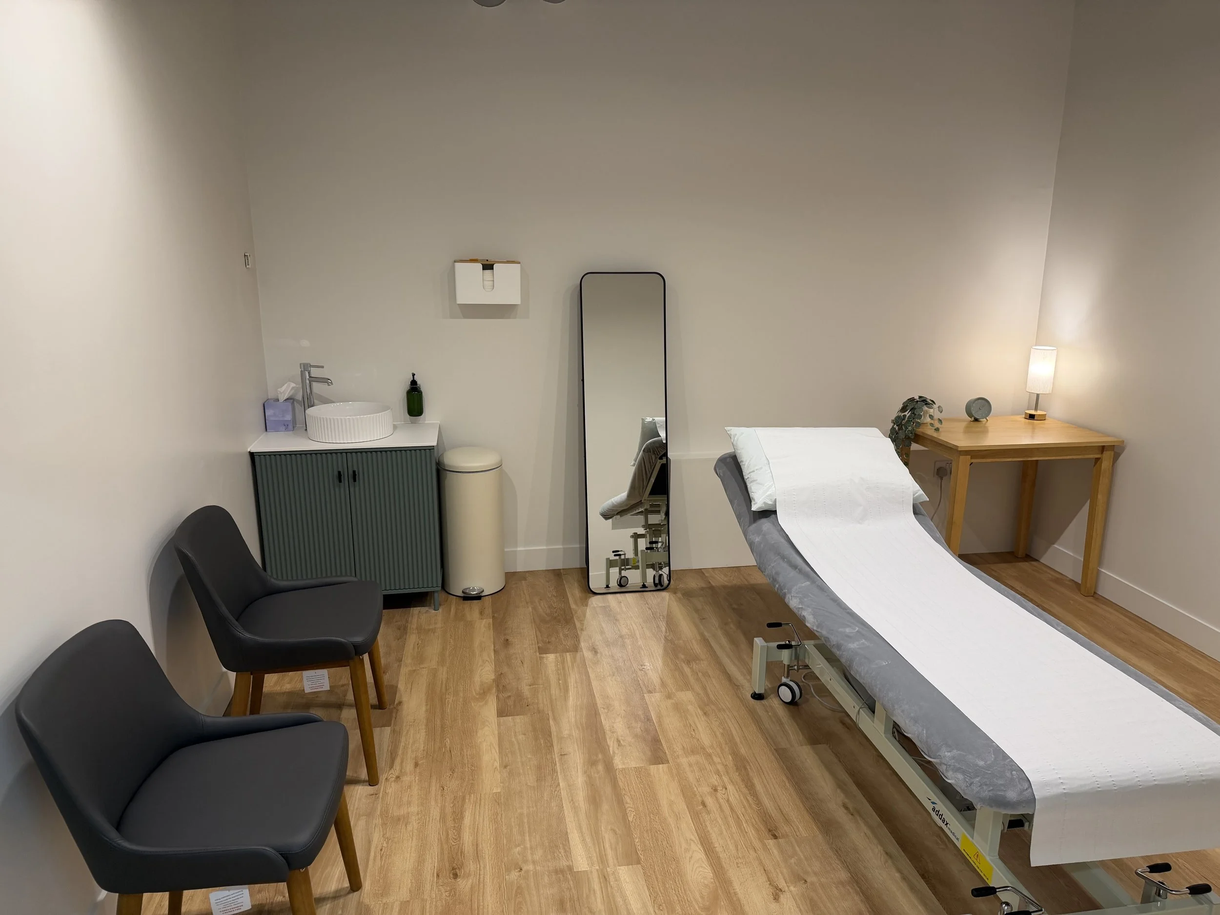 A medical examination room with a bed, chairs, a mirror, sink, lamp, and medical supplies.