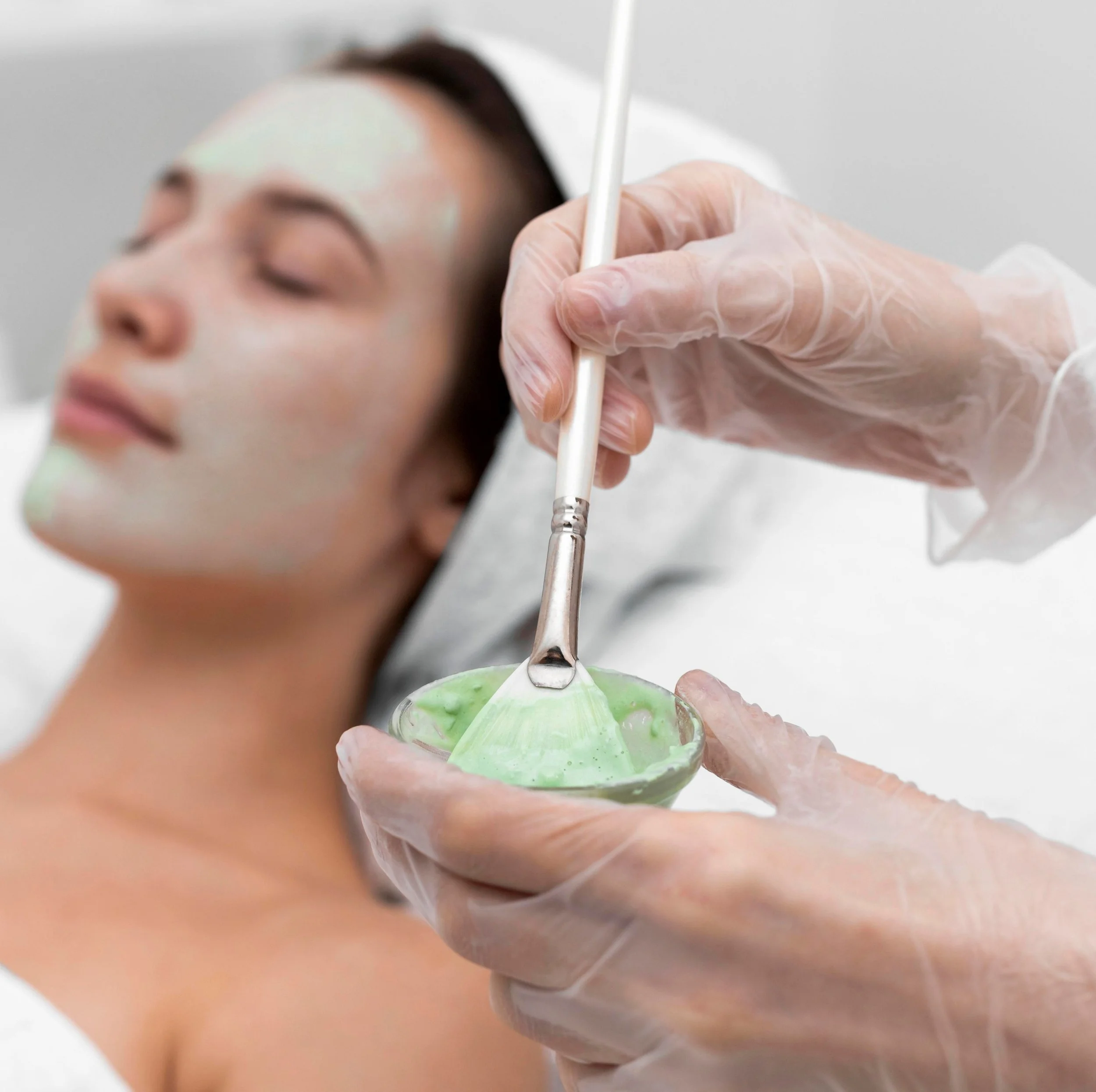 Close-up of a medical professional's gloved hand using a brush to apply a treatment to a person's face for skincare purposes.