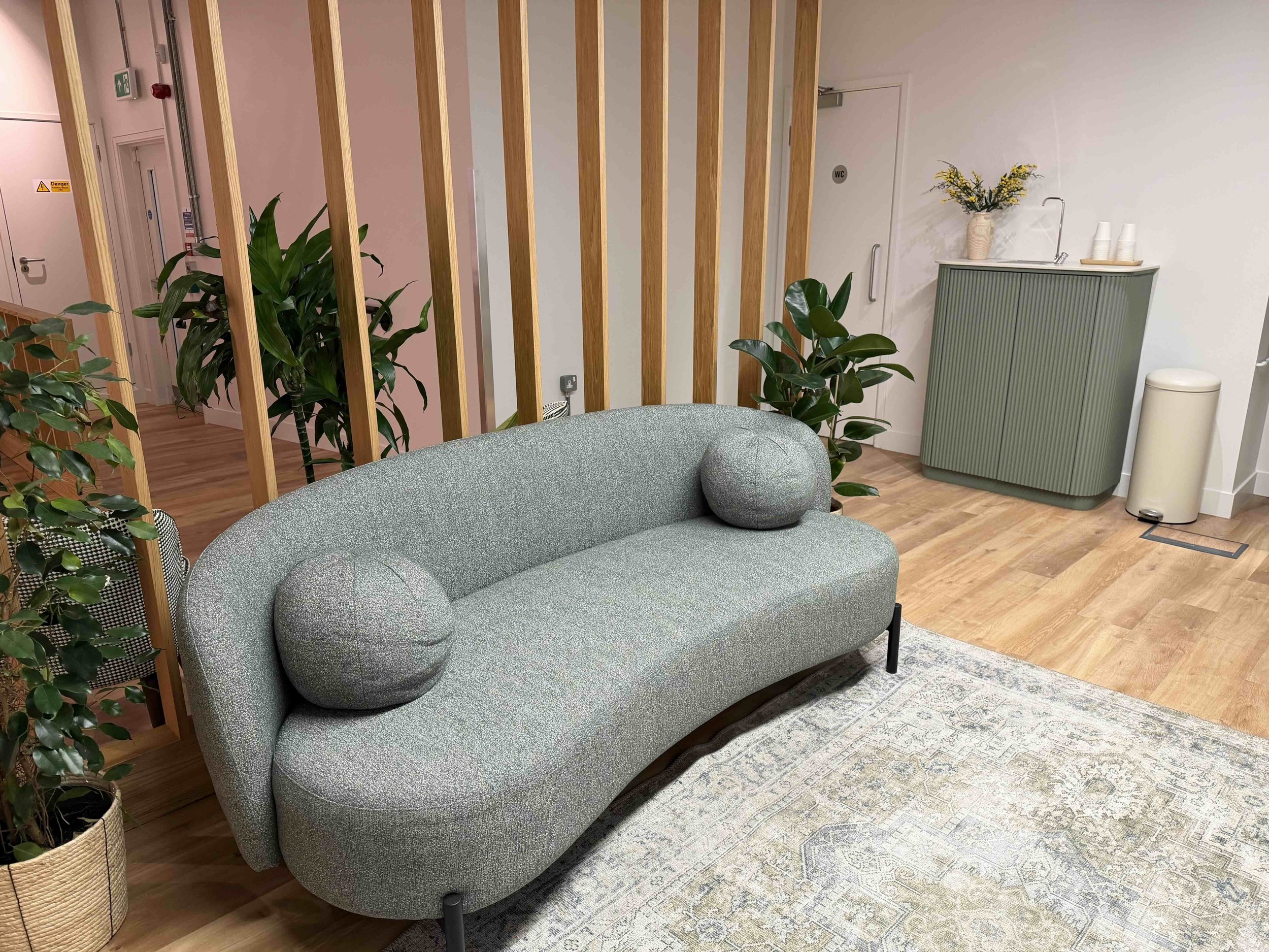 Interior of a modern room with a gray upholstered sofa, green plants, a wooden slat partition, a light green cabinet with a vase and cups, a white trash bin, and a sink with a tall faucet.