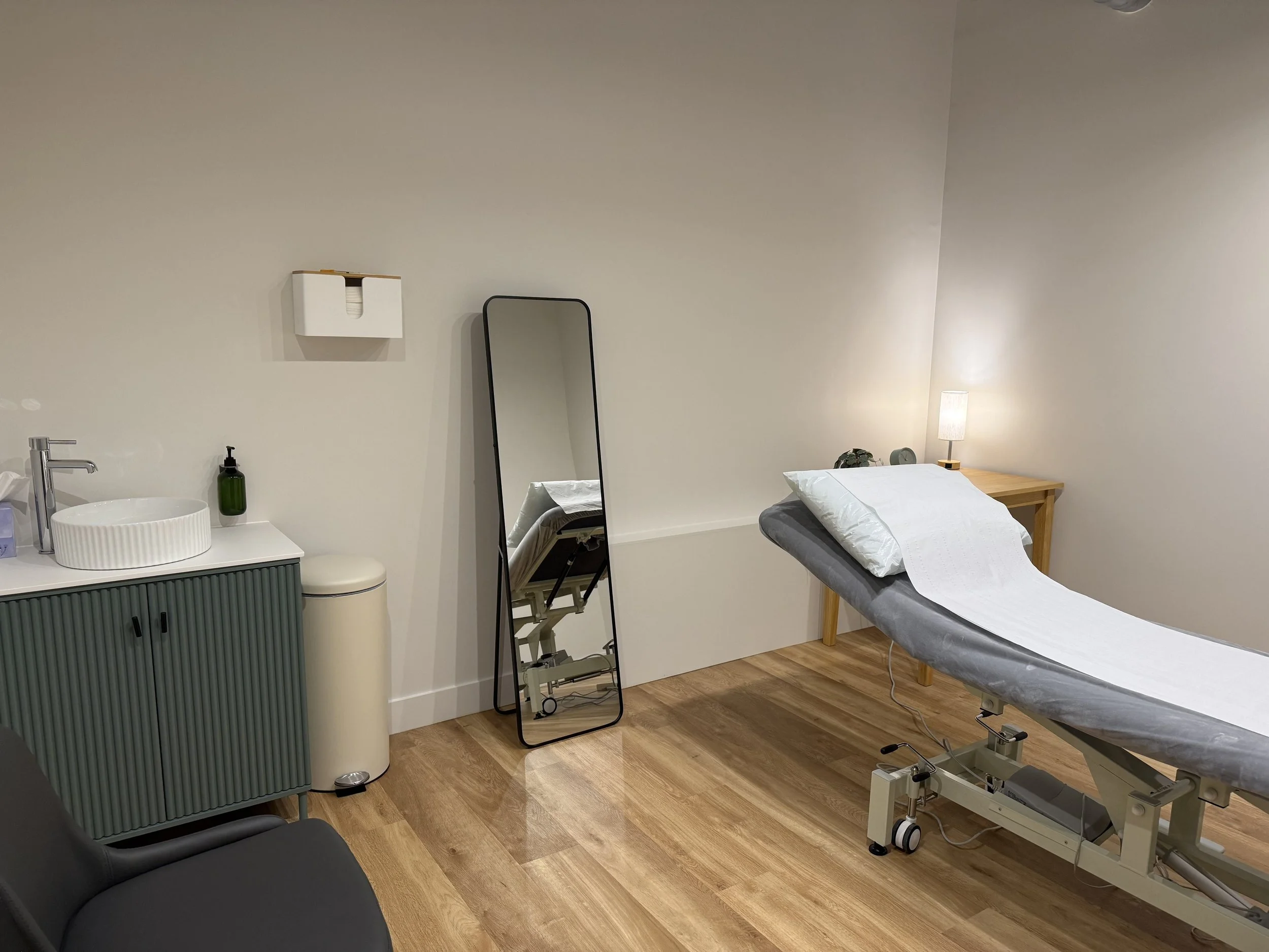 A doctor's examination room with an adjustable hospital bed, a large mirror, a lamp, a small cabinet, a sink, and a chair.
