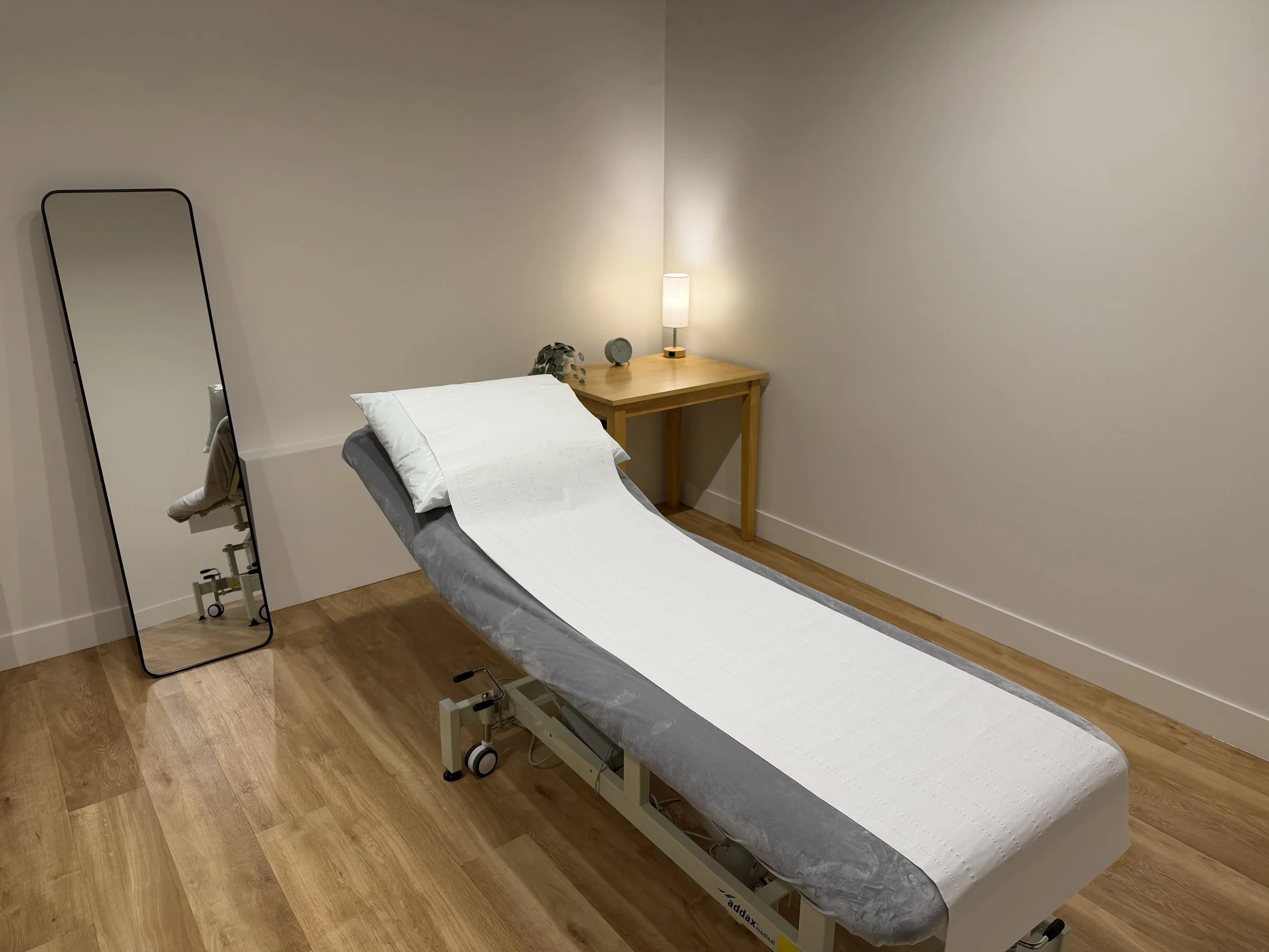 Medical examination room with a patient bed, a full-length mirror, a table with a lamp and plant, and wood flooring.