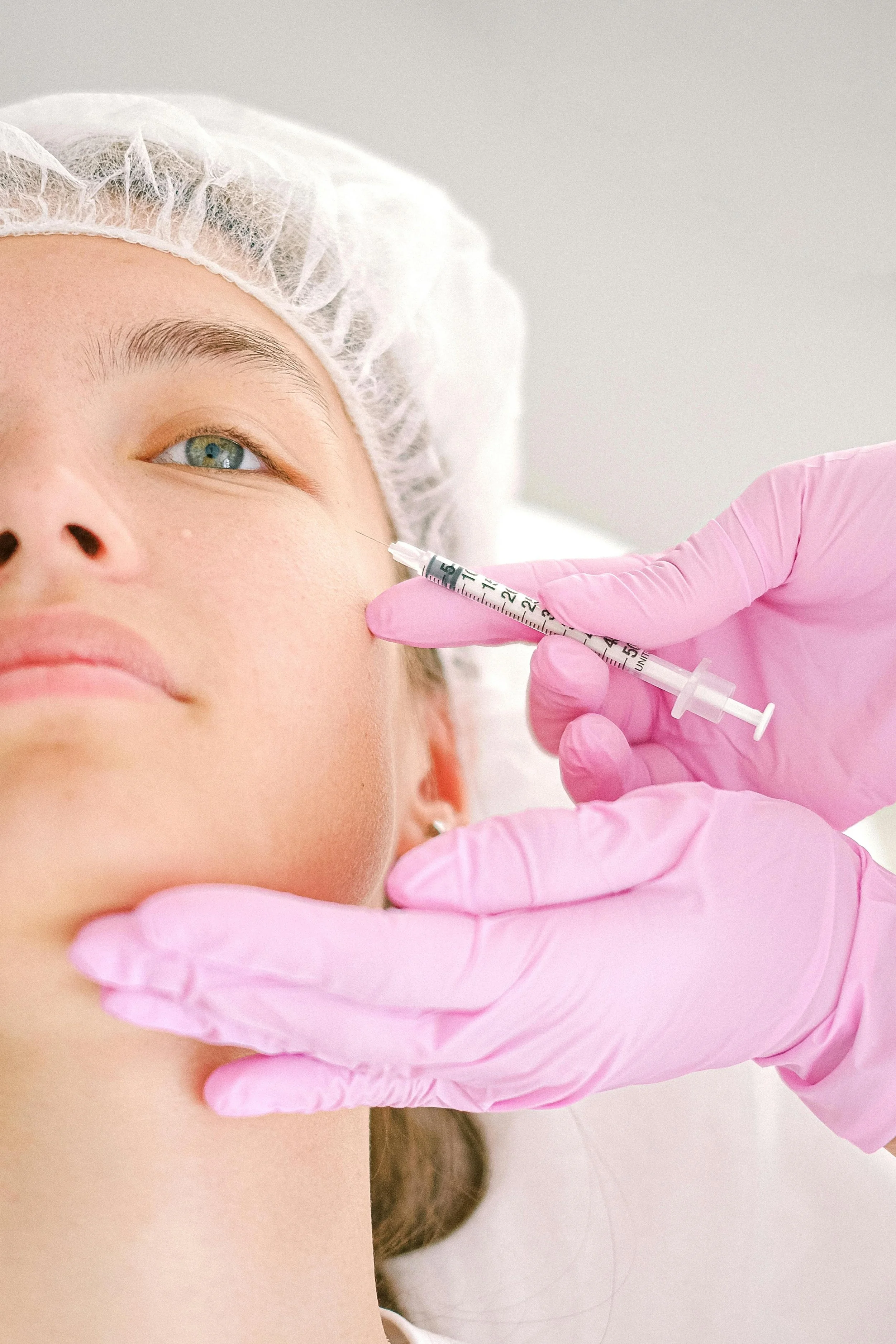 A medical professional administering a facial injection near a patient's eye using a syringe, wearing pink gloves.