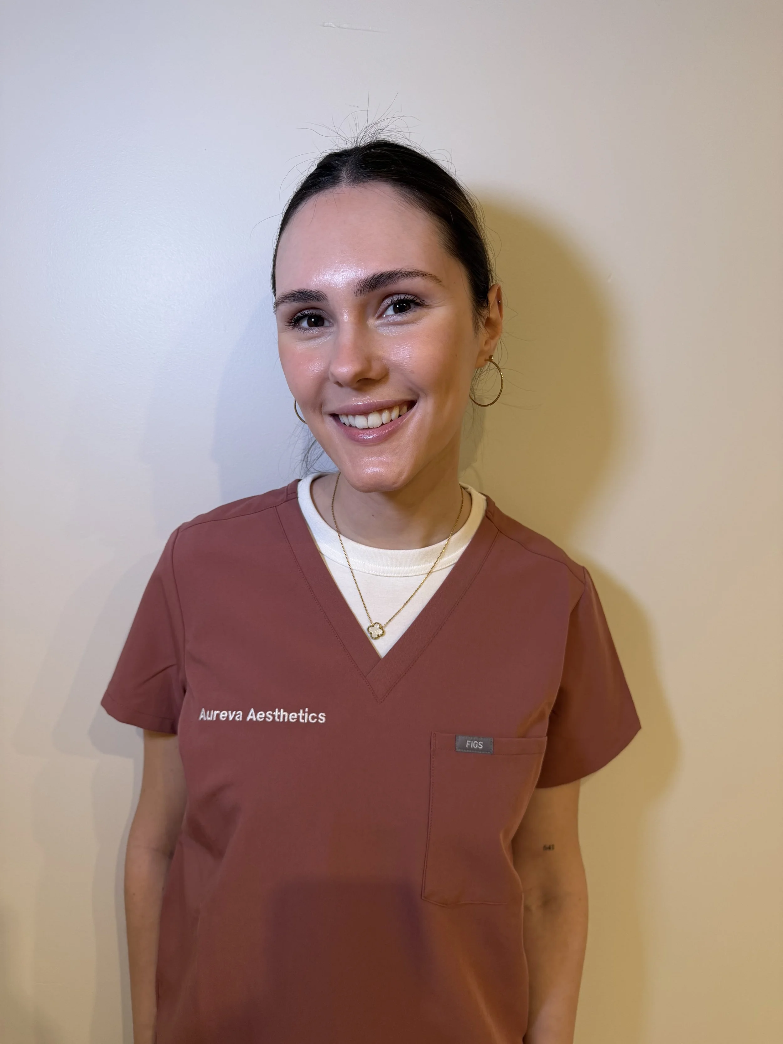 Aureva's founder, Beatrice, wearing a burgundy medical uniform with white text 'Aureva Aesthetics' on the left side. She has dark hair, gold hoop earrings, and is smiling, standing against a plain, light-colored wall.
