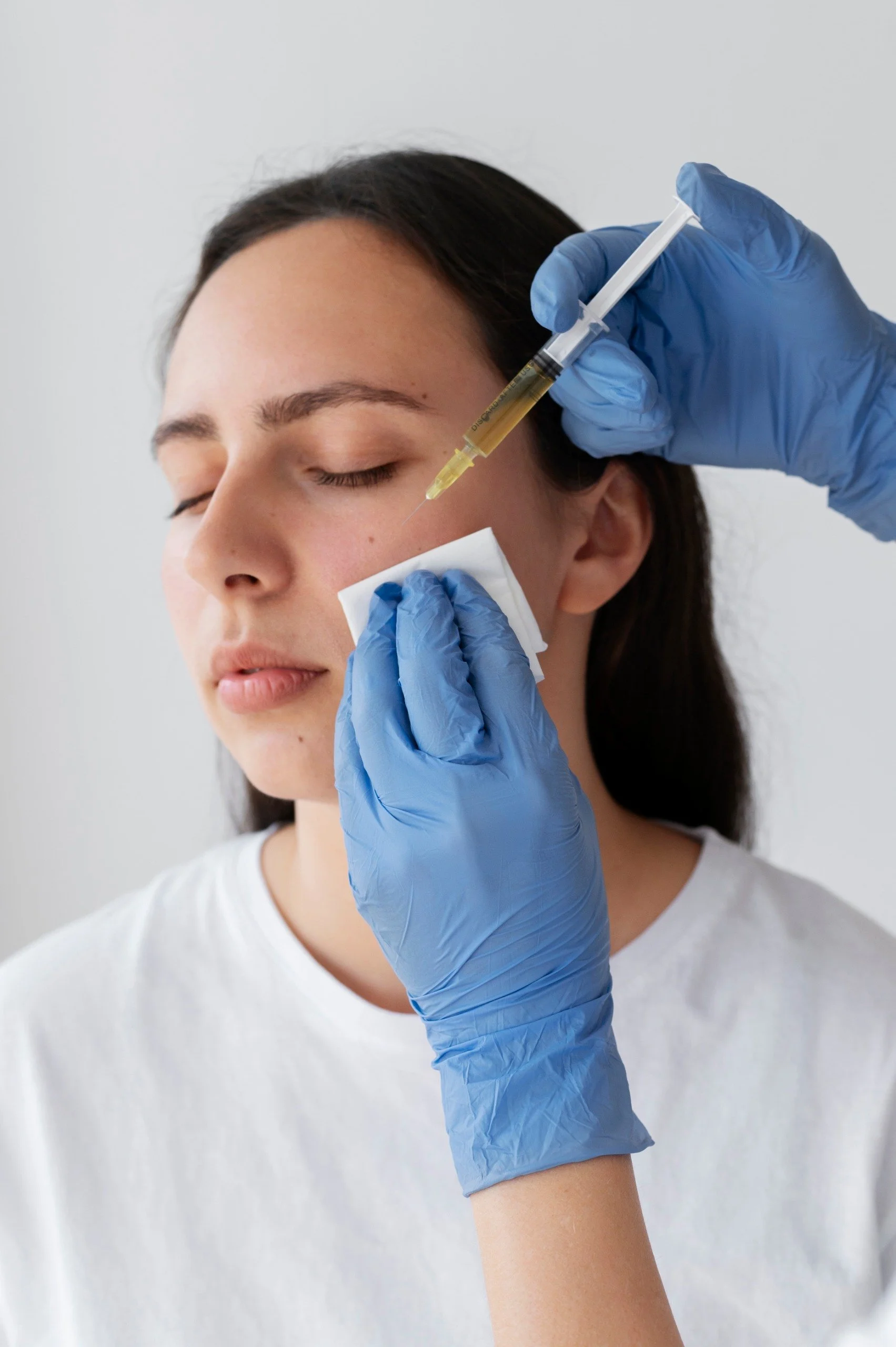 A woman with dark hair and closed eyes receiving an injection in the cheek from a healthcare professional wearing blue gloves, while holding a cotton swab against the person's face.