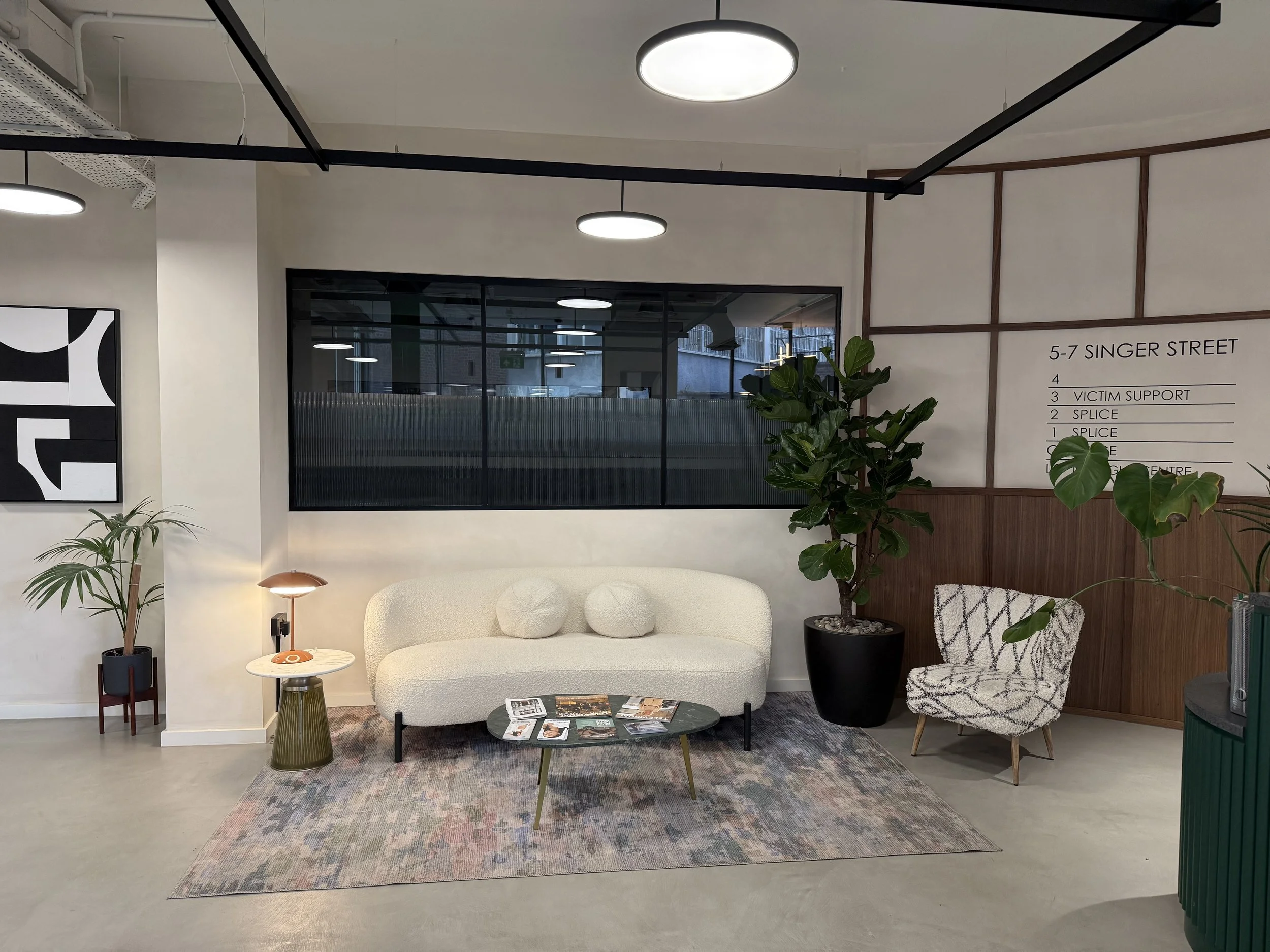 Interior of a modern waiting or lounge area with a white sofa, a small round coffee table with magazines, a patterned armchair, a potted plant, and decorative artwork on the wall. There are ceiling lights and a large window in the background.