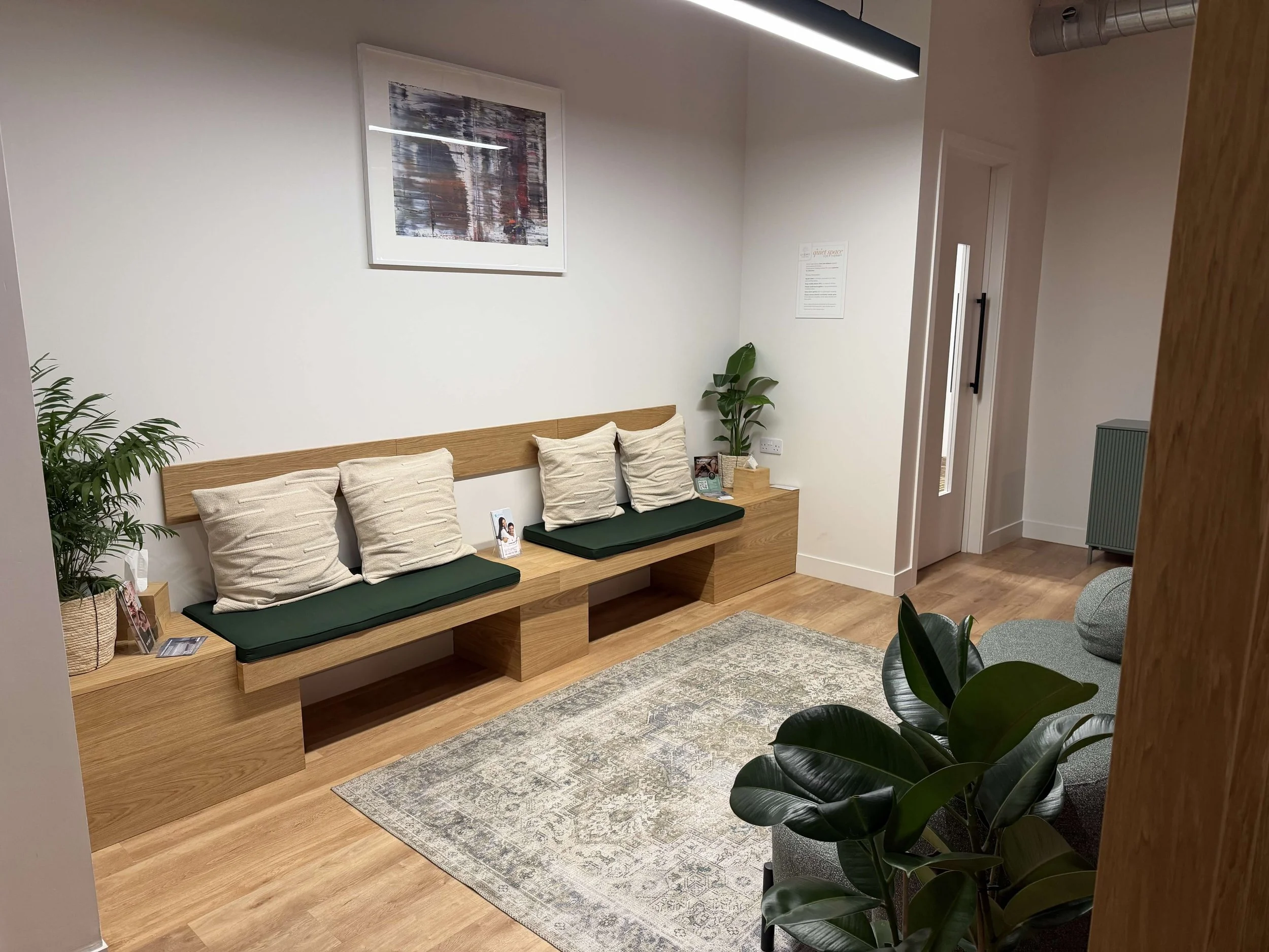 A cozy waiting room with a wooden bench that has green cushions and beige pillows, a potted plant, a patterned rug, and an abstract painting on the white wall. There is a small table with magazines and a tissue box, and a doorway to the right.