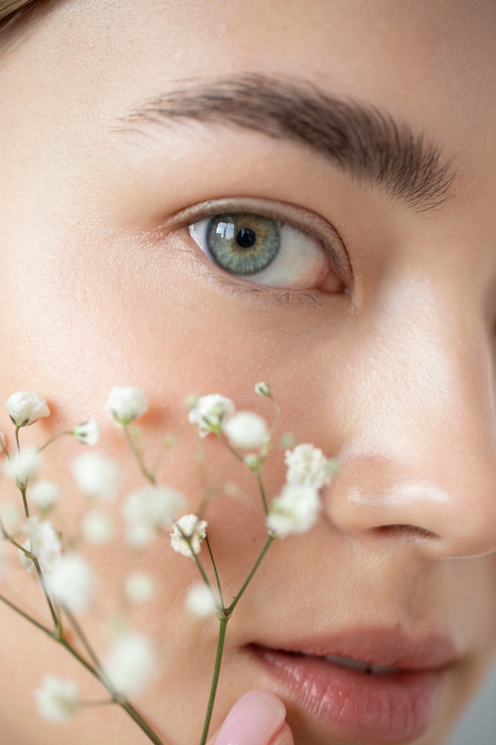 Close-up of a woman's face with an emphasis on her green eye, soft skin, and full lips, holding delicate white flowers near her face.