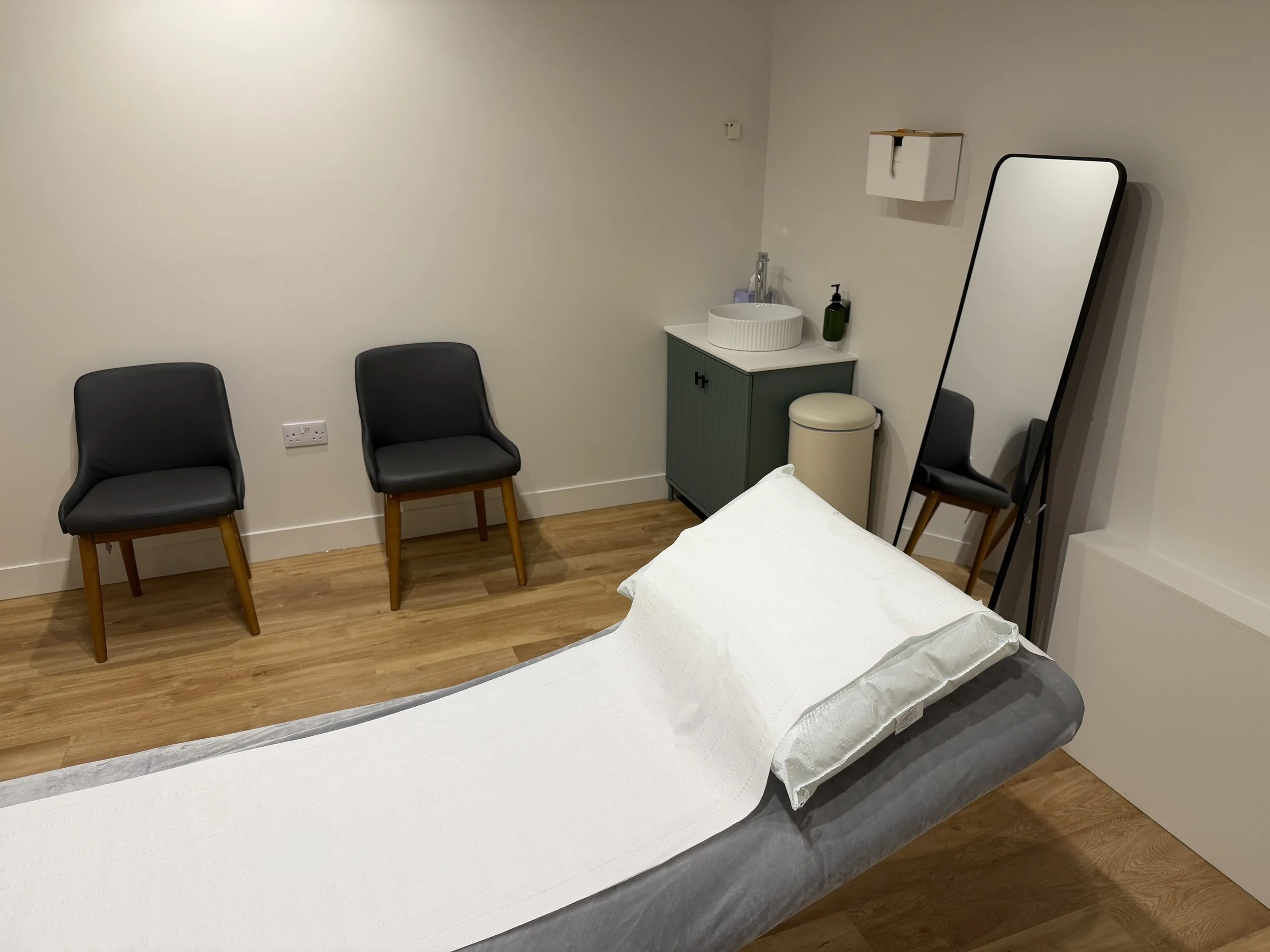 A medical examining room with a patient bed covered in white paper, two black chairs against the wall, a small cabinet with a sink and soap dispenser, a mirror, and a trash can.
