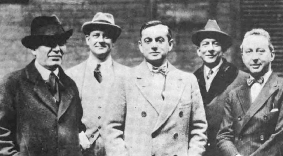 Black and white photo of five men standing close together, dressed in suits and hats, smiling.