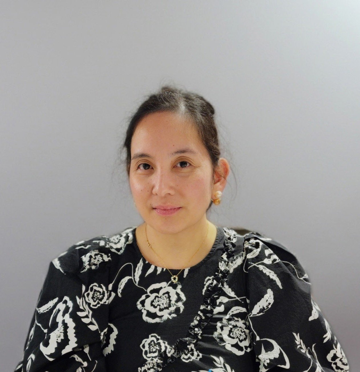Portrait of a woman with dark hair in a bun, wearing a floral black and white dress, pearl earrings, and a gold necklace, sitting against a plain gray background.