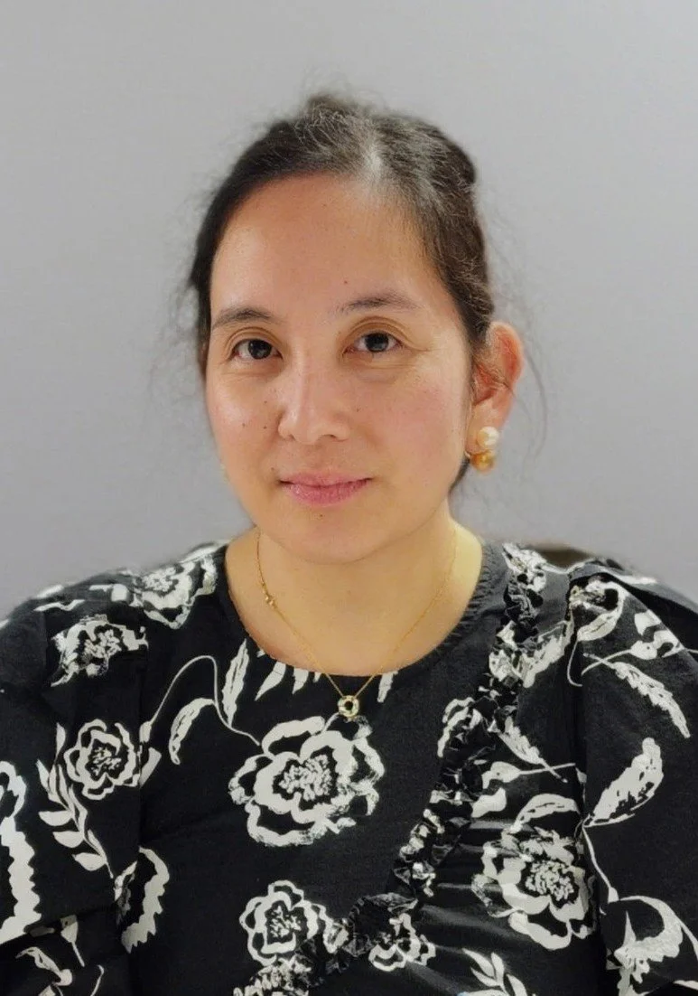 A woman with dark hair tied back, wearing a black dress with white floral pattern, gold jewelry including a necklace, earrings, and a ring, sitting against a plain gray background.