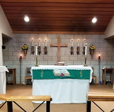 Church altar with green cloth, wooden cross, candles, flowers, and a Bible against a gray brick wall.