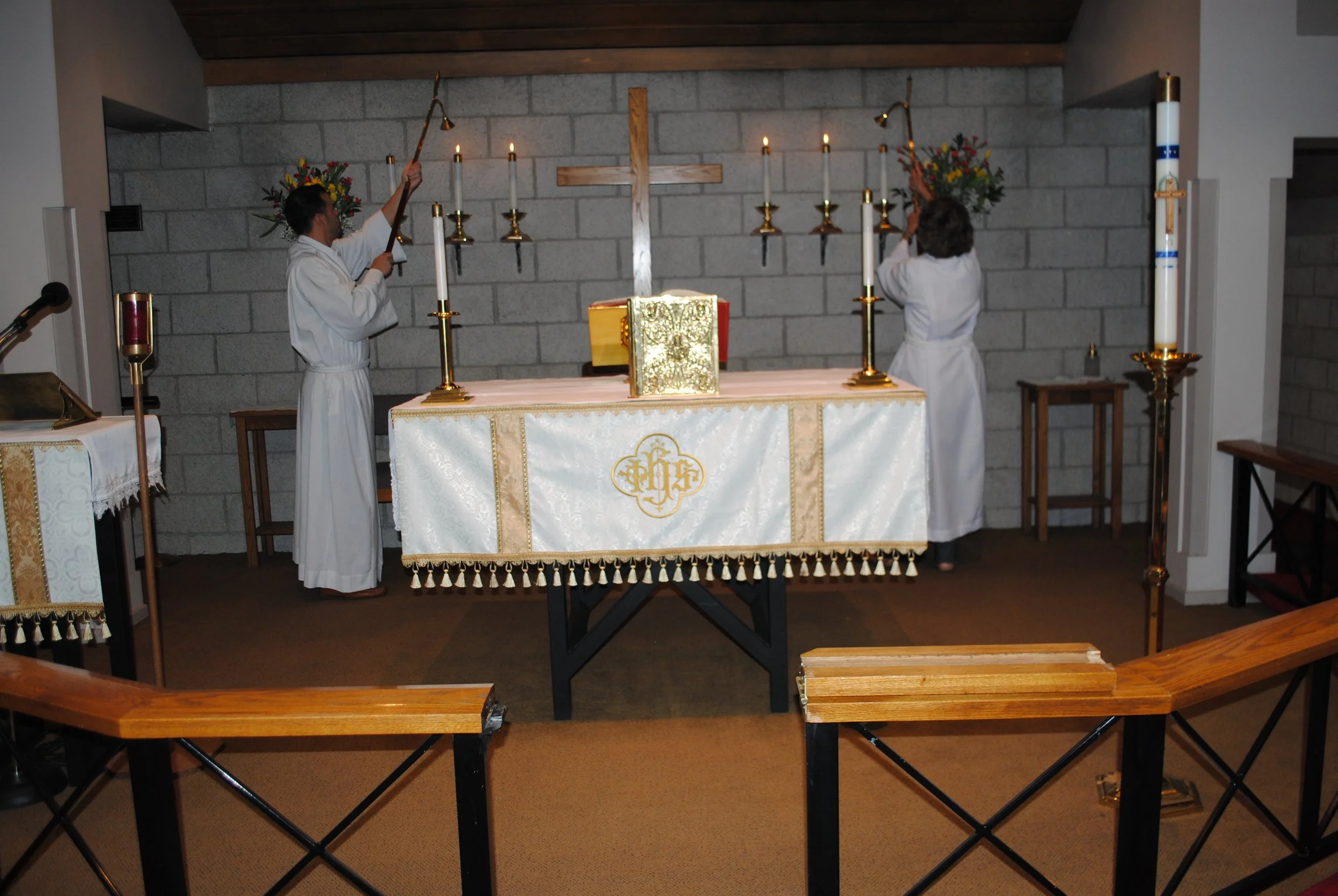 Two altar servers in white robes lighting candles on a church altar with a wooden cross and flowers. The altar is covered with a white cloth with gold embroidered details.