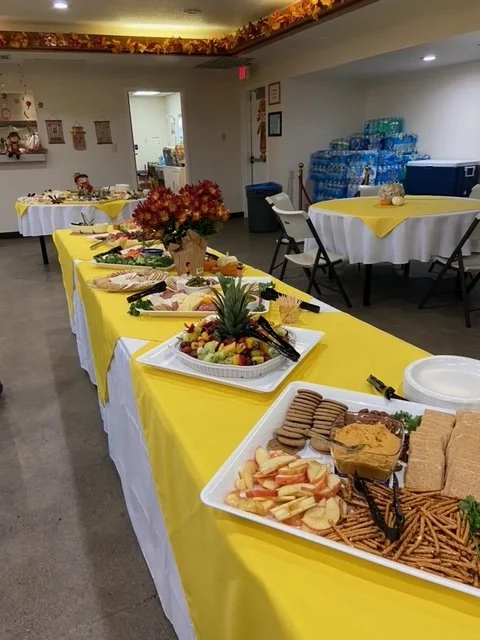 Buffet table with yellow tablecloths set up with various snacks, fruits, and desserts in a decorated room for a celebration or party.