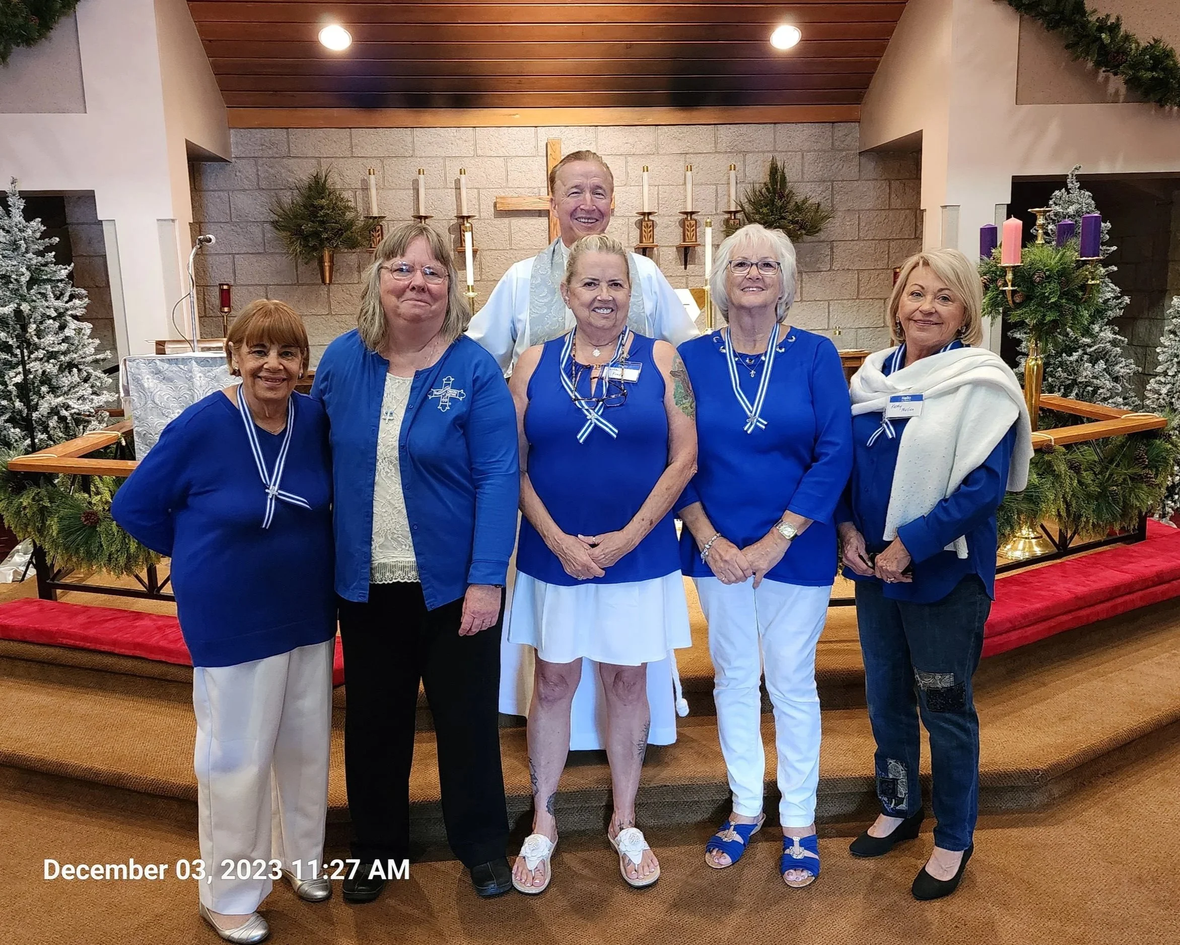 Group of six women and one man standing inside a church with a cross and candles in the background, dressed in blue shirts, some with medals and ribbons, celebrating Christmas.