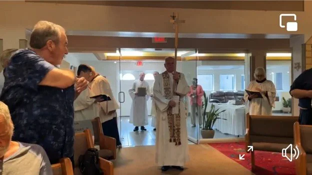 A priest holding a cross leads a group of clergy and congregation in a church service or prayer. Some individuals are holding prayer books, and others are standing or sitting in the church.