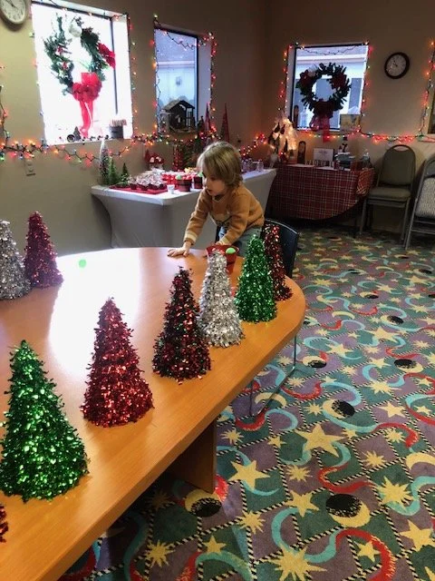 A young child decorating a table with small, glittery Christmas trees in red, green, silver, and gold. The room has Christmas wreaths, holiday lights, and festive decorations, with a table set for Christmas and a colorful star-patterned carpet.