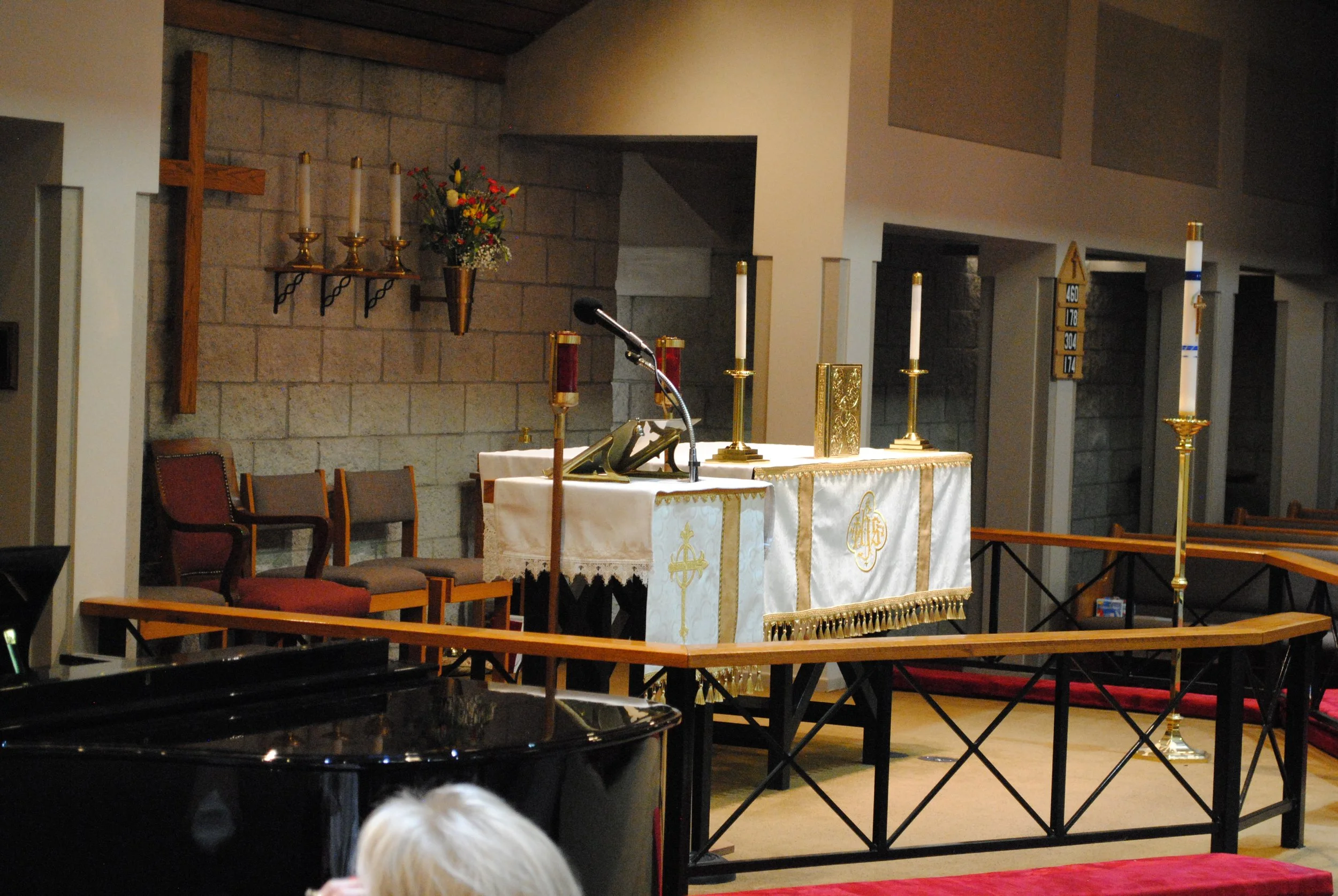 Interior of a church altar with a white cloth, gold candlesticks with candles, and a floral arrangement. Chairs are arranged beside the altar, and a wooden cross is mounted on the wall to the left.