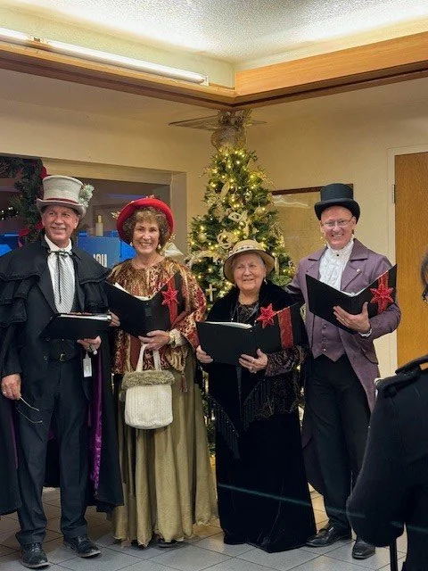Four people dressed in vintage, Victorian-style costumes standing in front of a decorated Christmas tree, holding songbooks and appearing to perform holiday carols.