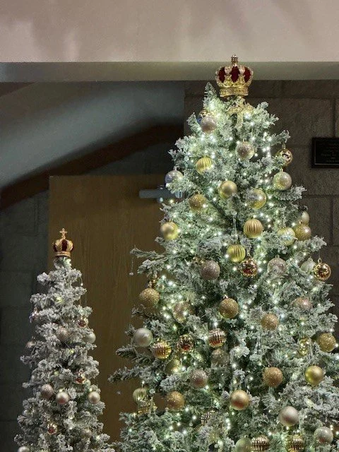 Two decorated Christmas trees, one larger and one smaller, with gold, silver, and pink ornaments and topped with crowns, in a room with a wooden door and gray walls.