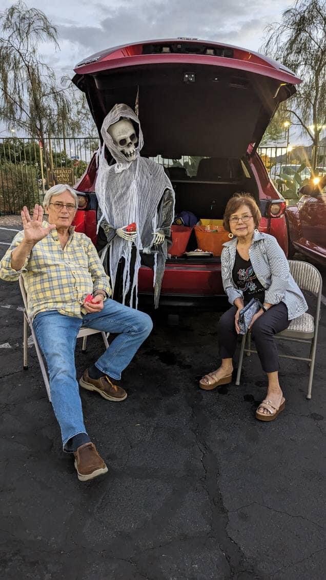 Two people sitting on chairs in front of an open car trunk with a Halloween skeleton decoration dressed in a hooded robe.