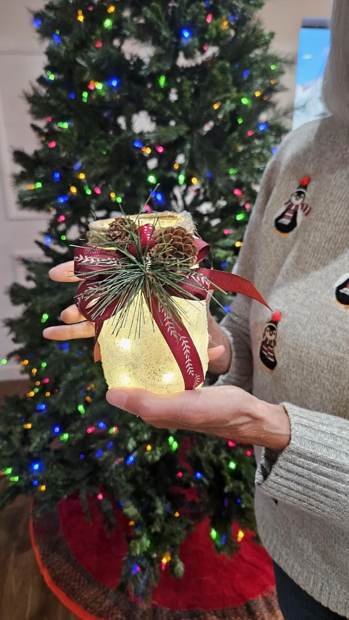 A person holding a small decorative Christmas lantern decorated with a red ribbon, pine cones, and pine needles, with a Christmas tree decorated with multicolored lights in the background.