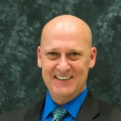 A bald man in a suit and tie smiling at the camera, with a dark, blurred background.