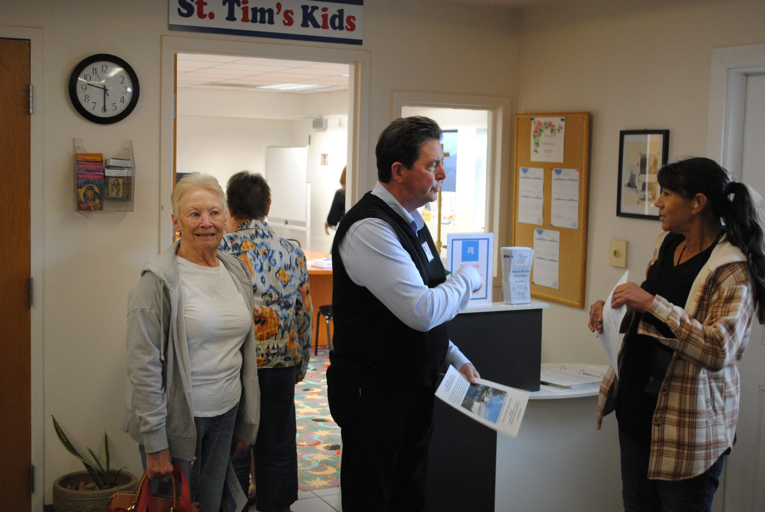 A woman with short blonde hair and a light gray hoodie standing in line at a reception desk, with others including a woman holding papers and a man in a vest and light blue shirt talking to a staff member in a busy indoor setting, under a sign readin