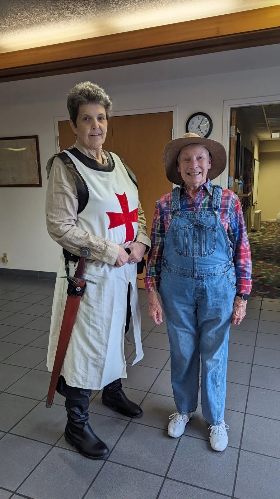 Two smiling elderly women dressed in costumes, one as a medieval knight with a red cross on a white tunic, the other as a farmer with a straw hat, plaid shirt, and denim overalls, standing indoors in a hallway.