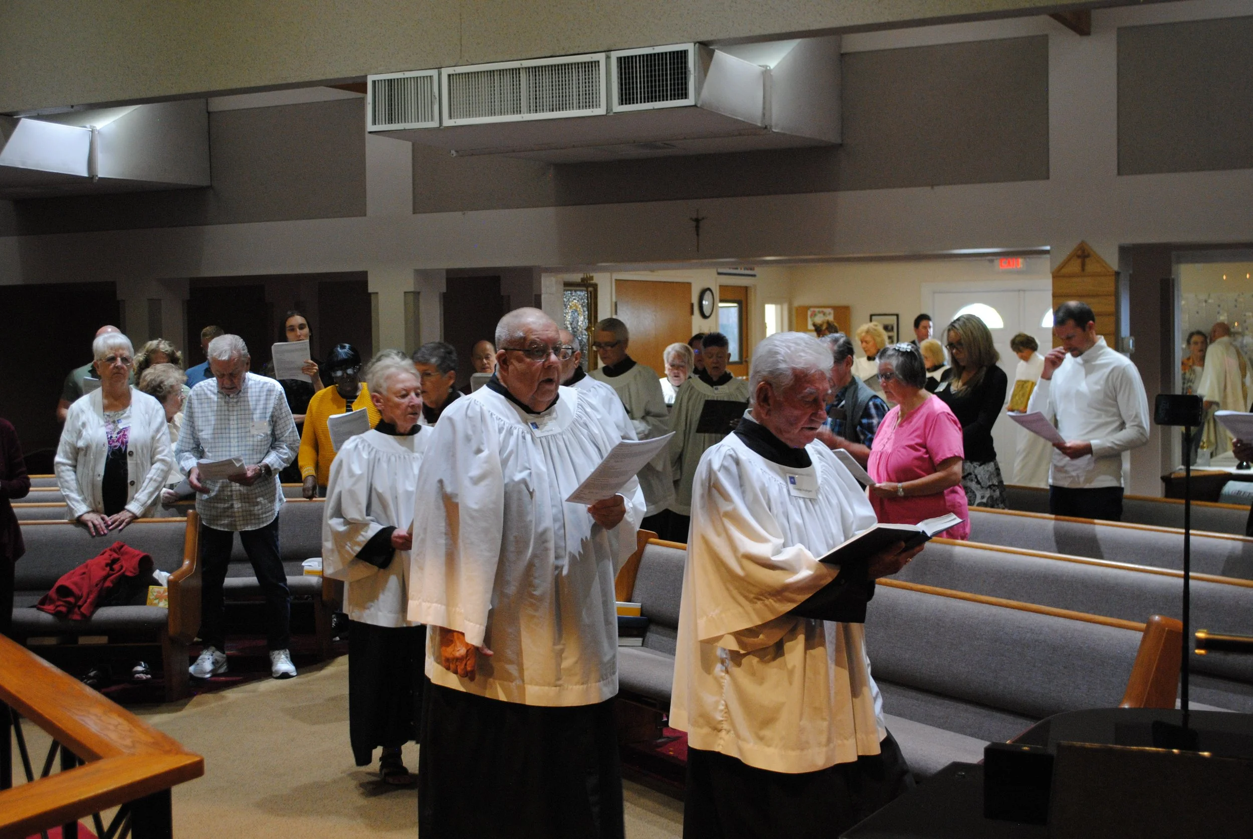 People gathered in a church, participating in a prayer or hymn, some wearing choir robes, with pews and church interior visible.