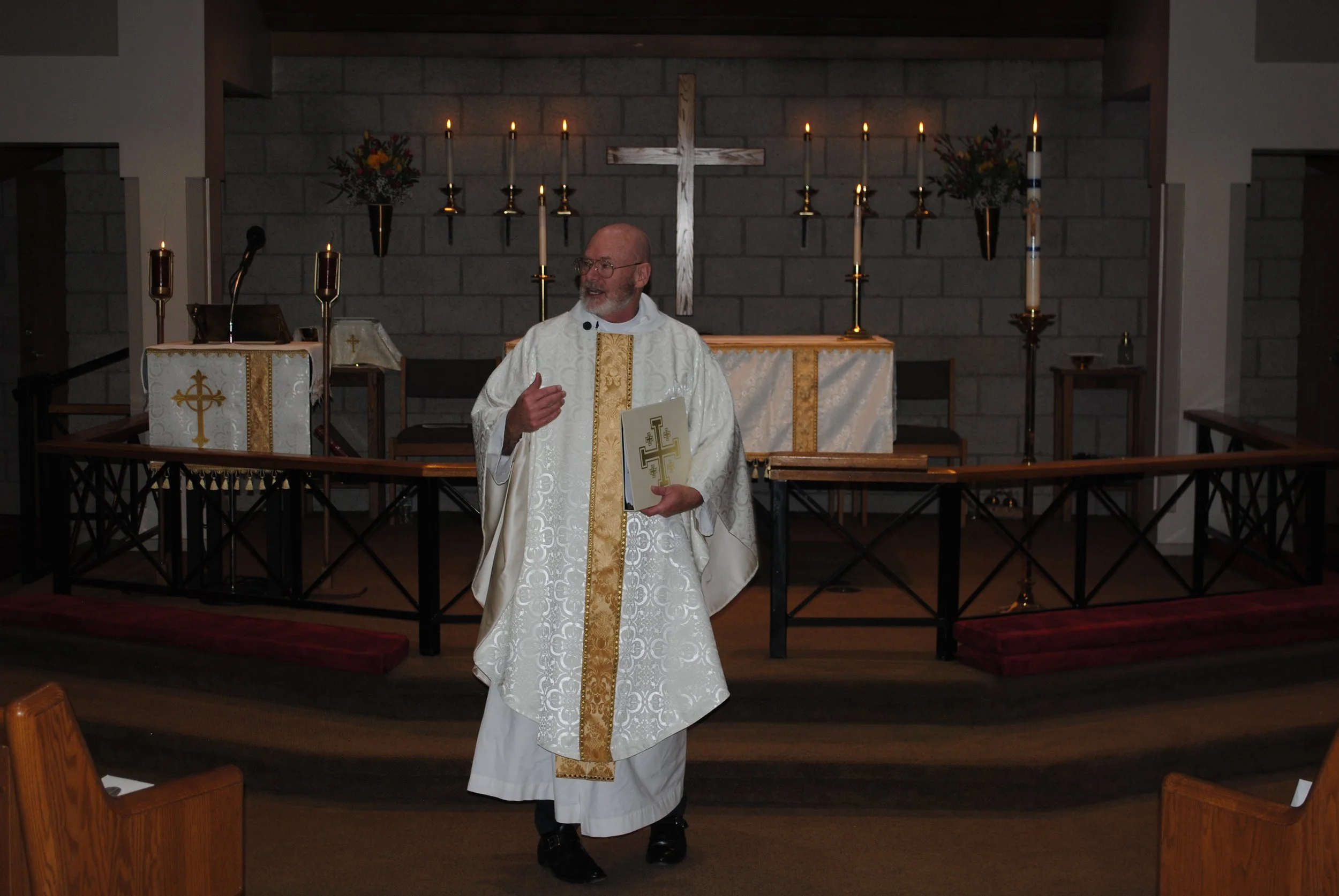 A priest in white liturgical vestments standing in a church, holding a booklet with a cross symbol, in front of an altar with lit candles, flowers, and a cross in the background.