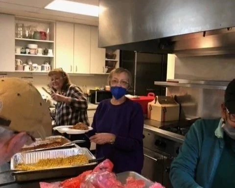 People serving food in a kitchen, with a woman smiling and wearing a blue mask, and a man in a blue shirt and mask.