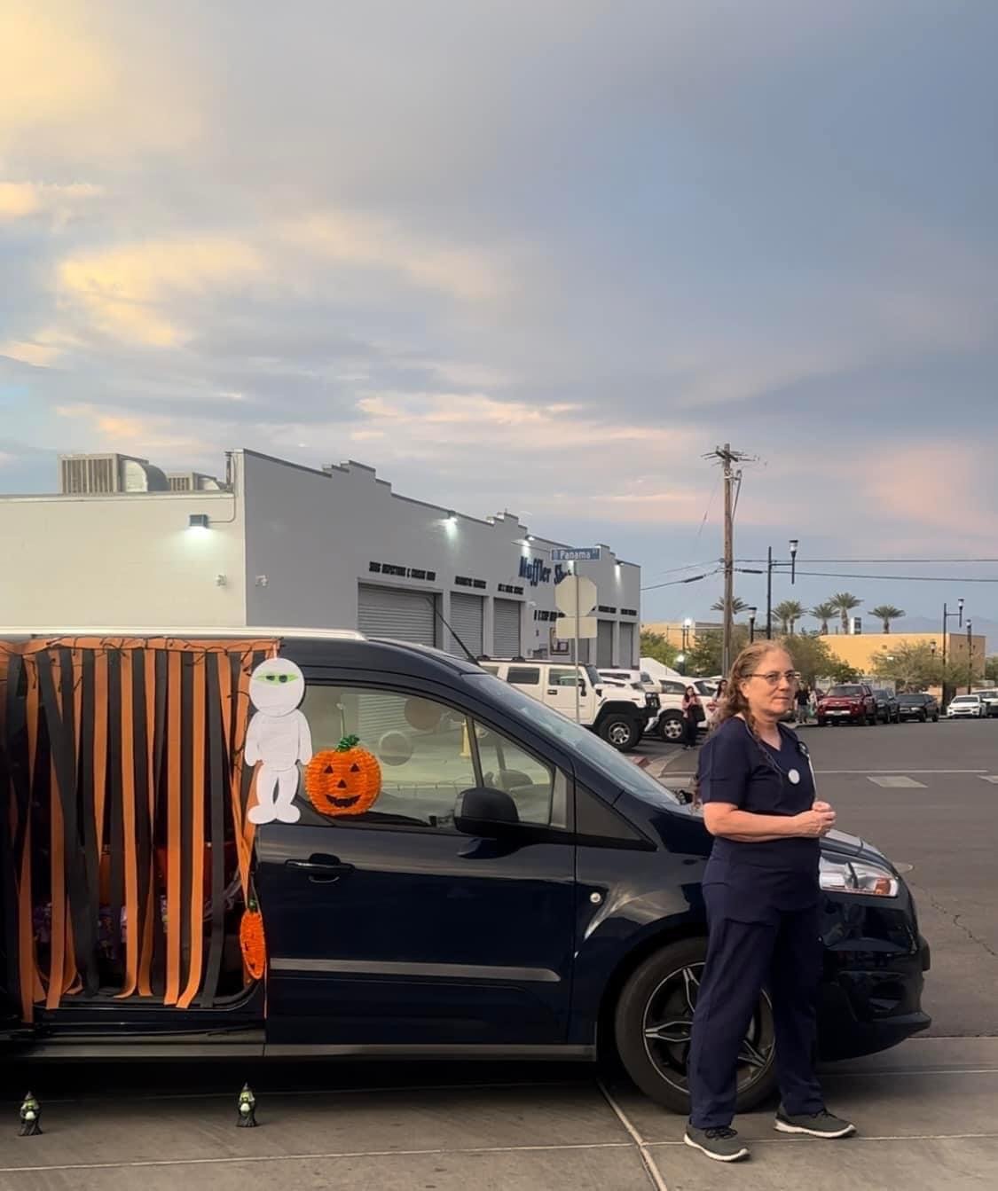 A woman in navy scrubs standing next to a decorated vehicle with a Halloween theme, featuring paper streamers, a paper cutout of a white character, and a paper pumpkin with a face, in a parking lot outside a shopping center at dusk.