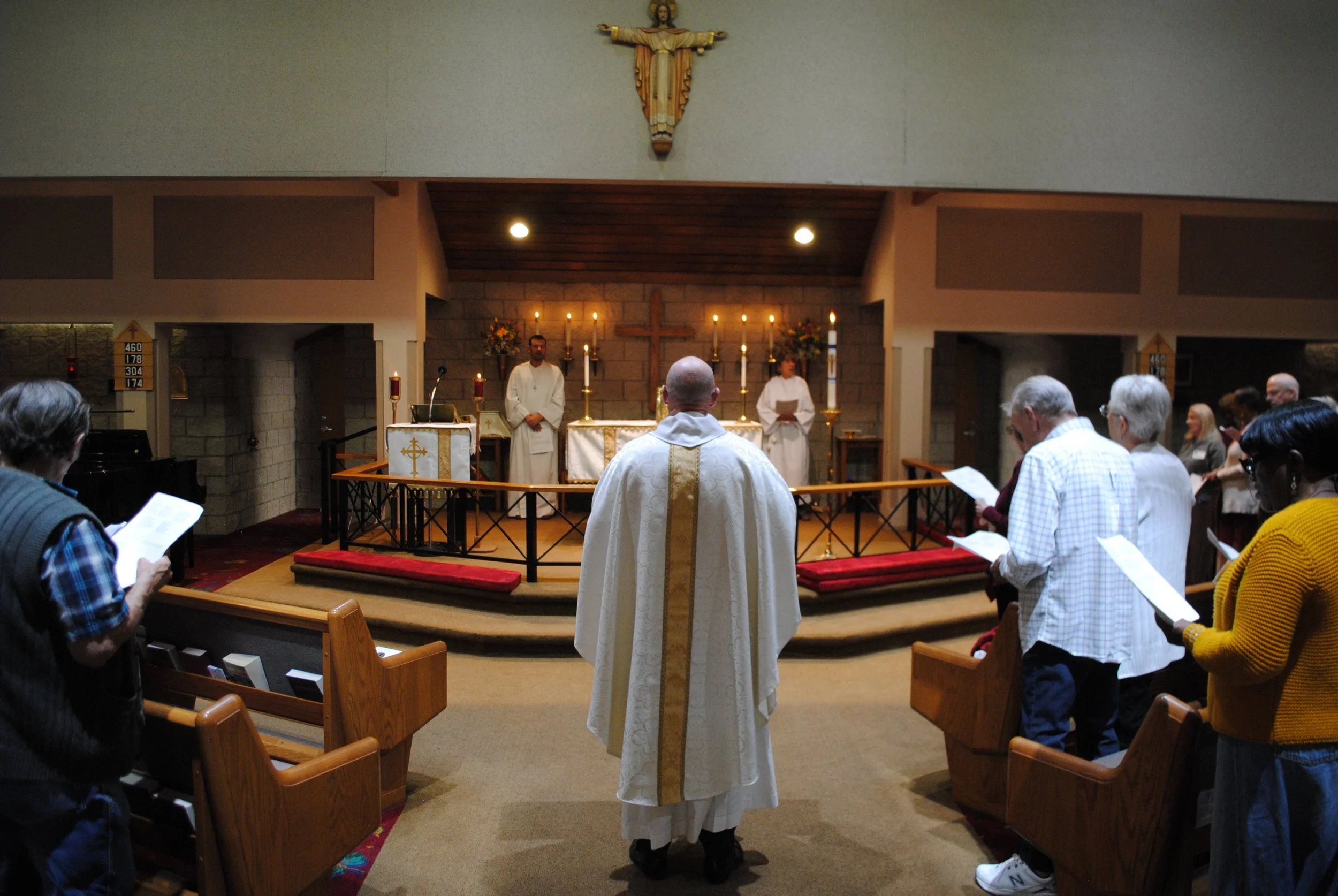 Inside a church during a service, a priest in white robes stands at the altar with his back to the congregation. The altar is decorated with candles and flowers, with a large cross and statue of Jesus on the wall behind. The congregation is praying a