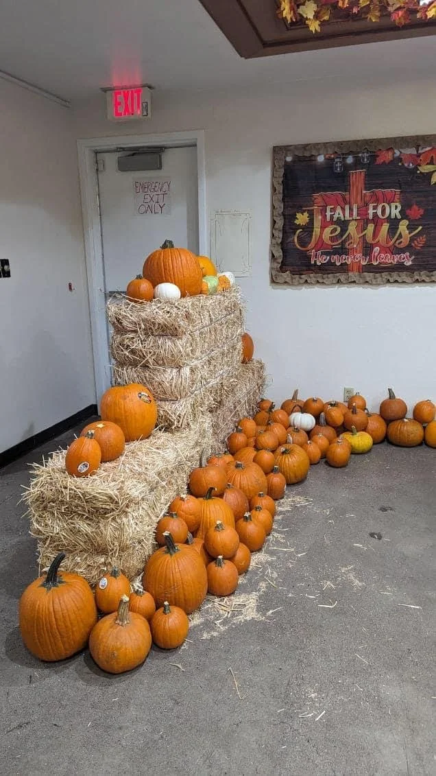 Display of pumpkins arranged on hay bales and floor against a wall with fall-themed decorations and a sign that reads "Fall for Jesus."