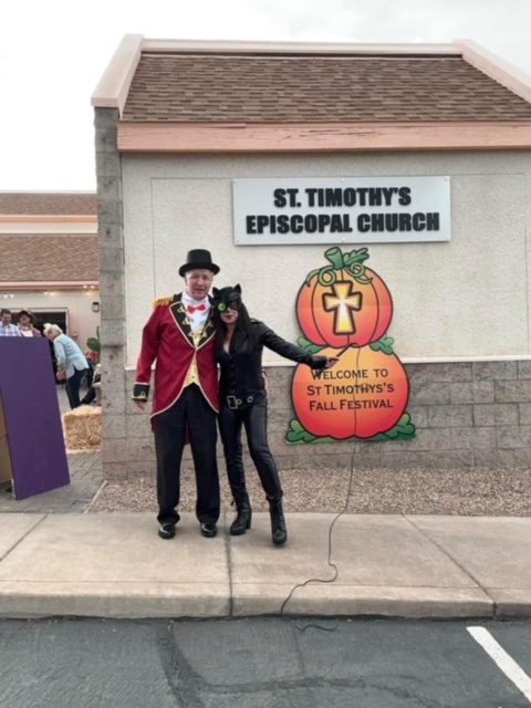 Two people in costumes stand in front of a sign for St. Timothy's Episcopal Church at a fall festival. One is dressed as a circus ringmaster, the other as a black cat wearing a mask.
