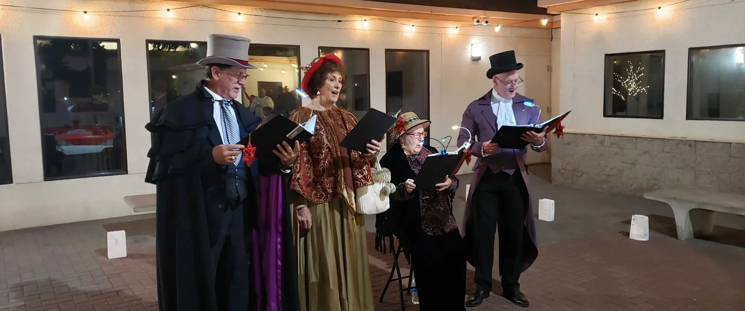 A group of five people dressed in vintage holiday costumes singing carols outdoors at night, with Christmas lights in the background.