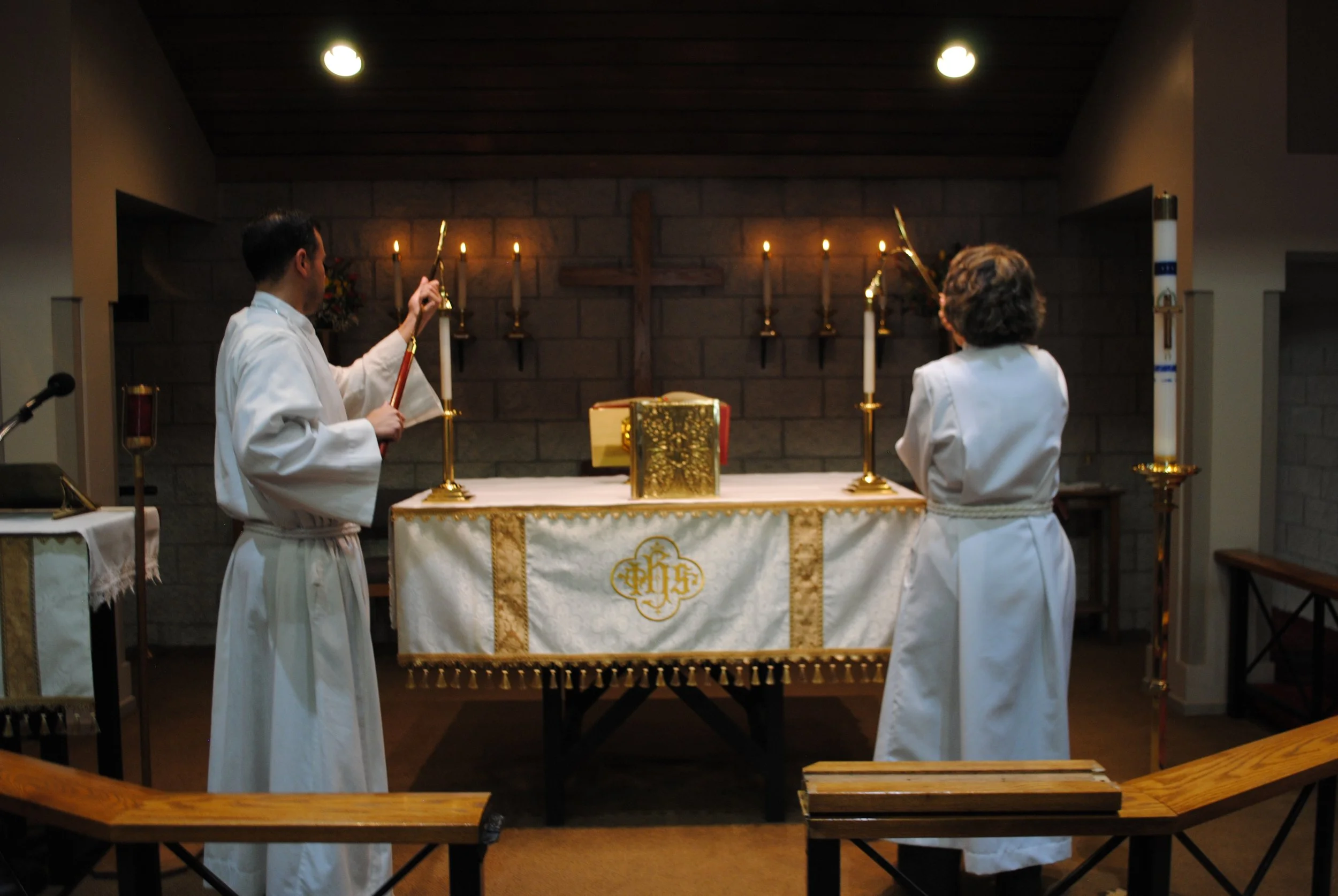 Two priests in white robes standing at an altar in a church, lighting candles during a religious service, with a wooden cross behind them and a brick wall as the background.