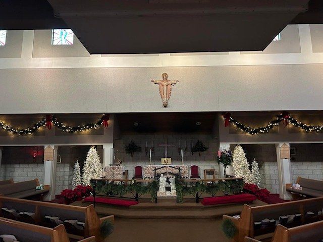 The interior of a church decorated for Christmas with garlands, poinsettias, and Christmas trees, featuring an altar with a cross and Christmas wreaths, and a wall with a crucifix and a statue of Jesus.