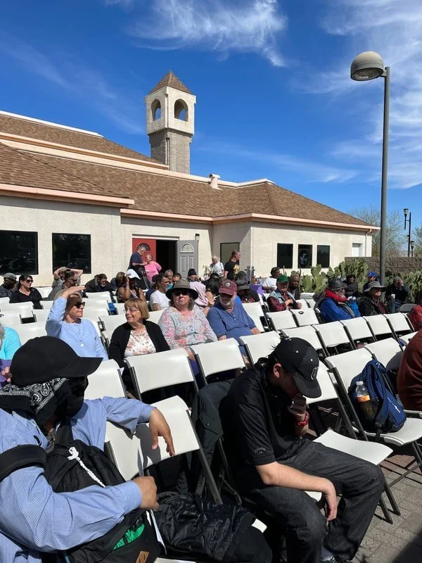 People seated outdoors in front of a building with a clock tower, under a blue sky with some clouds.