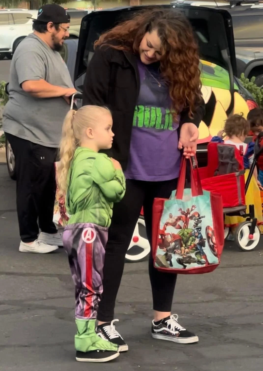 A woman with curly red hair, wearing a black jacket, purple shirt, and black pants, is holding bags featuring Marvel characters. A young girl wearing a green hoodie, Marvel-themed pants, and green shoes is standing nearby, looking at her. They are ou