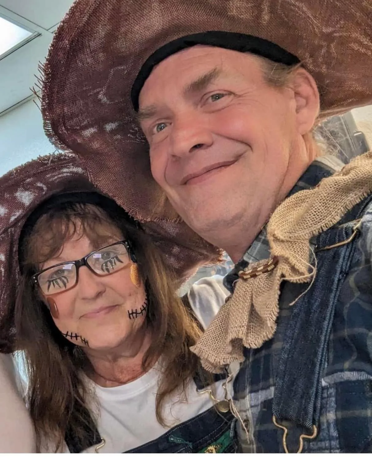 Two people dressed in Halloween costumes, wearing large straw hats. The woman has face paint resembling a skeleton with glasses and curly hair. The man has a friendly smile, long hair, and is wearing a plaid shirt with a scarf around his neck.