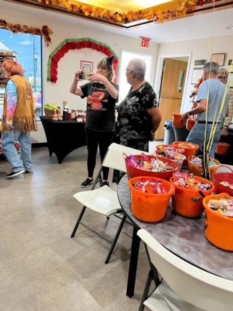 Indoor room decorated with autumn leaves, featuring a table with orange bowls filled with snacks, and several people including two women in the center, one taking a photo, at a gathering or party.