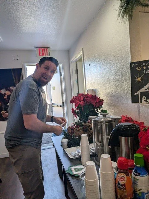 Man standing at a beverage table with coffee supplies, cups, and Christmas decorations inside a building.