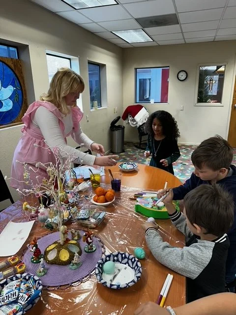 Children and a woman decorated Easter eggs at a table with Easter decorations, eggs, and craft supplies in a room with colorful windows and a patterned carpet.