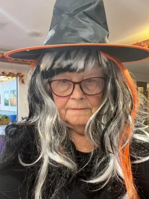 Older woman wearing a witch's hat with a black and orange color scheme, long gray and black wig, glasses, and a black shirt.