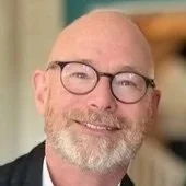 A smiling bald man with a beard and glasses, wearing a suit jacket