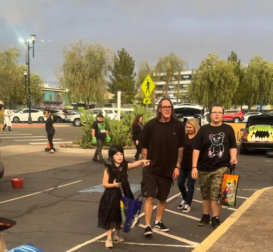 A group of people walking in a parking lot, including a woman, a little girl pointing, and a young man carrying a gift bag, with cars and a street scene in the background.